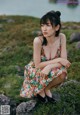 A woman in a floral dress sitting on a rock by the water.